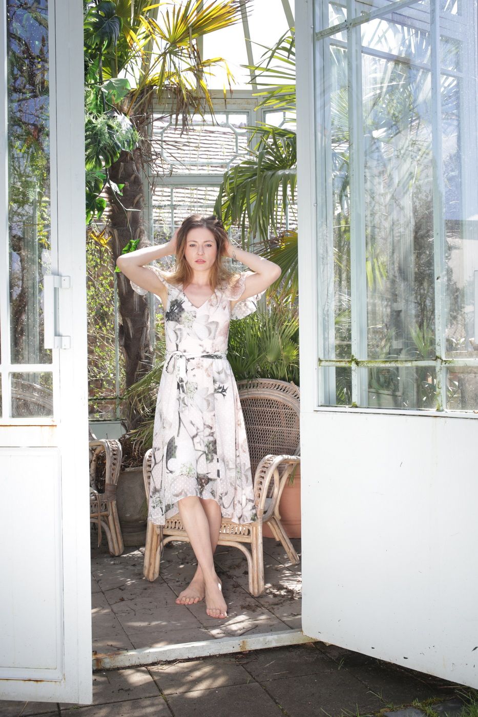 Irina le Fey im geblümten Kleid auf einem Korbstuhl im Gewächshaus